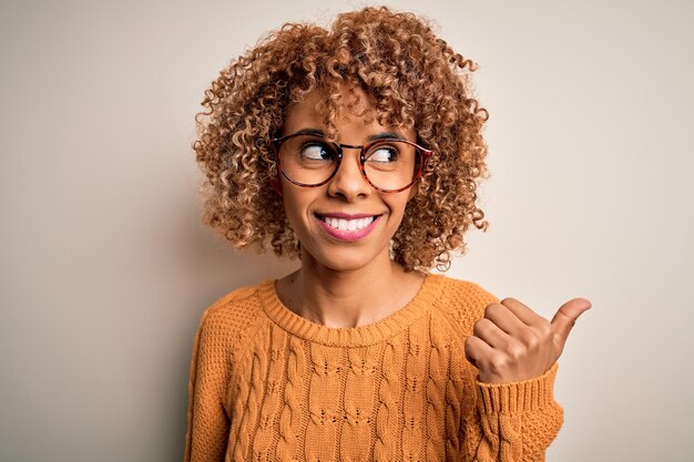 白い背景にカジュアルなセーターと眼鏡をかけた若いアフリカ系アメリカ人女性が幸せそうな顔をして微笑み、親指を立てて側面を指さしている