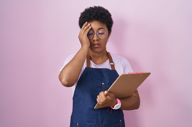 Young african american woman wearing professional waitress apron holding clipboard yawning tired covering half face, eye and mouth with hand. face hurts in pain.