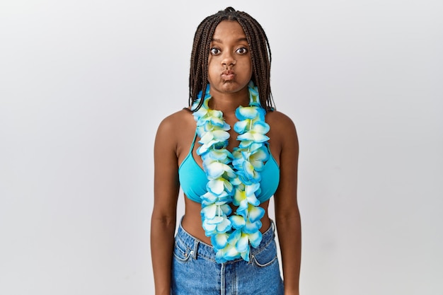 Free photo young african american woman with braids wearing bikini and hawaiian lei puffing cheeks with funny face. mouth inflated with air, crazy expression.