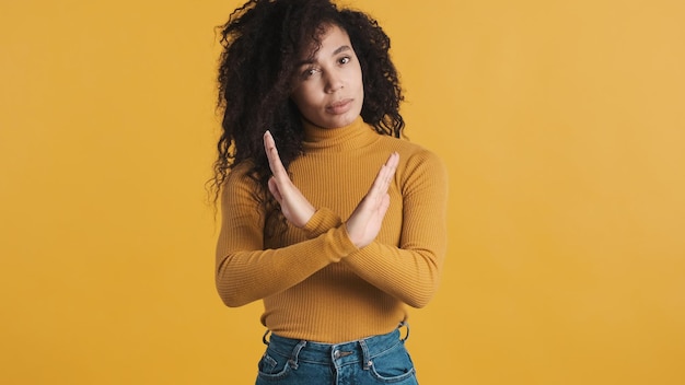 Giovane donna afroamericana con capelli scuri e soffici che sembra sicura di mantenere le mani incrociate che mostra no sulla fotocamera su sfondo colorato