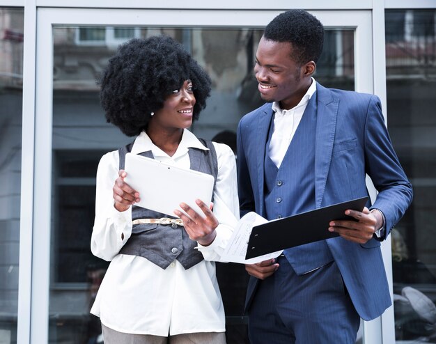 Young african businessman and businesswoman holding digital tablet and clipboard looking at each other