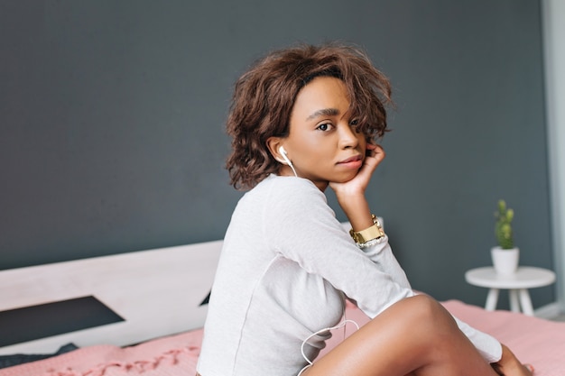 Free photo young african girl with short curly hair listening to music in earphones on bed, at home, sad mood. gray room. wearing light gray t-shirt with long sleeves, golden watches.