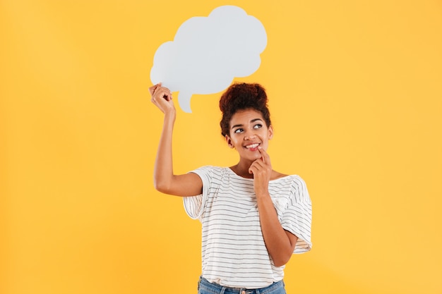 Free photo young african happy thoughtful lady looking up and holding blank speech bubble isolated over yellow