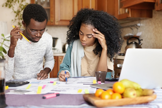 Young African husband and wife doing paperwork together at home, planning new purchase, calculating family expenses, sitting at kitchen table with laptop and calculator. Domestic budget and finances