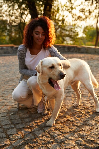 Young african lady in casual clothes sitting and hugging dog in park