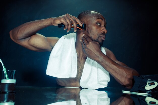 young african man in bedroom sitting in front of the mirror scratching his beard at home. Human emotions concept