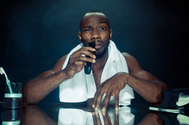 young african man in bedroom sitting in front of mirror scratching his beard at home