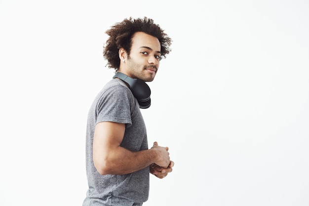 Young african man looking suspiciosly at camera standing in profile white wall with his wireless headphones neck.