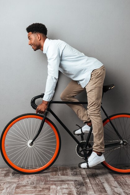 Молодой африканский человек стоя над серой стеной с велосипедом.