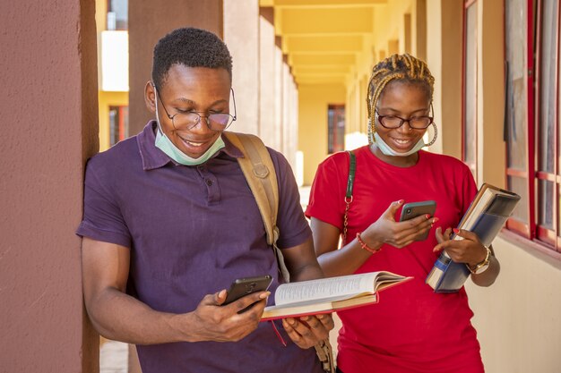 young african students wearing facemasks and holding books and phones at a campus
