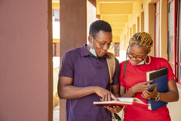 young african students wearing facemasks and holding books and phones at a campus