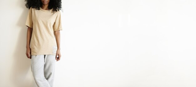 young African woman dressed in loose t-shirt and gray cotton pants standing cross-legged against white blank wall. Stylish dark-skinned student girl having rest indoors