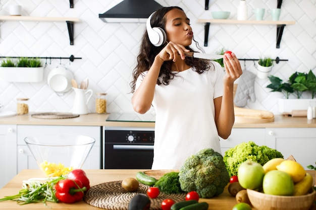 Young african woman emotional is listening to music via headphones and singing cuts a cherry tomato