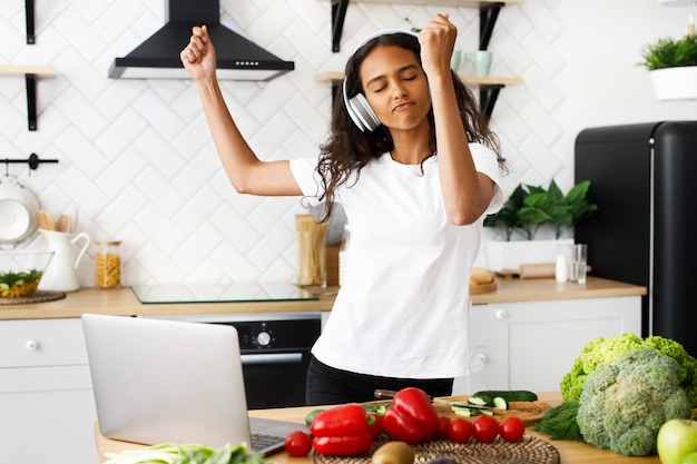 Young african woman is dancing and listening to music via headphones with closed eyes on the kitchen