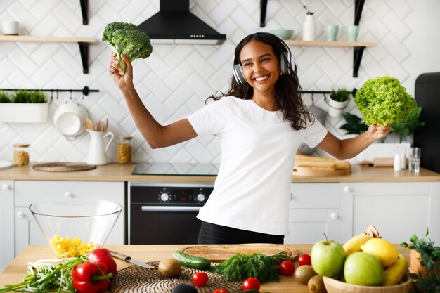 Young african woman is happy listening to music via headphones and holds a broccoli and salad