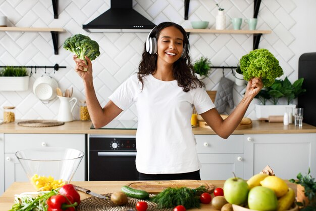 Young african woman is listening to music via headphones and holds a broccoli and salad