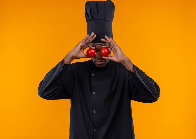 Free photo young afro-american cook in chef uniform closes eyes with tomatoes isolated on orange background with copy space