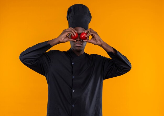 Young afro-american cook in chef uniform closes eyes with tomatoes on orange  with copy space