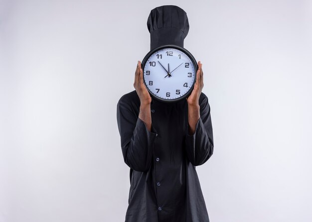 Young afro-american cook in chef uniform closes face with clock on white  with copy space