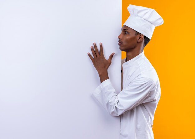 Молодой афро-американский повар в униформе шеф-повара стоит боком и кладет руку на белую стену, изолированную на оранжевом фоне с копией пространства
