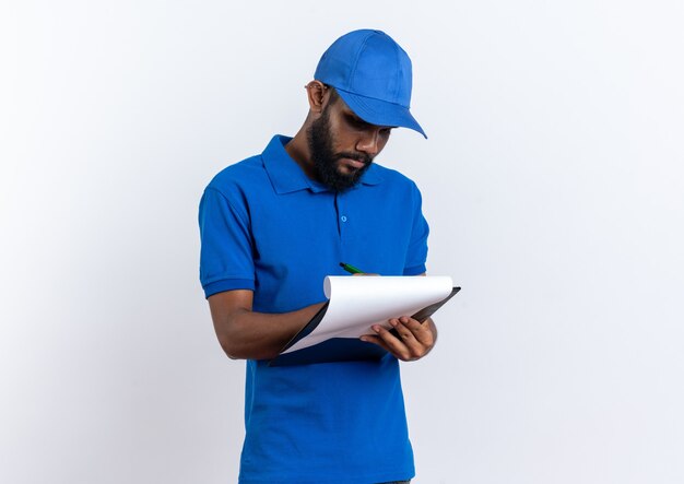 Young afro-american delivery man writing something on clipboard isolated on white wall with copy space