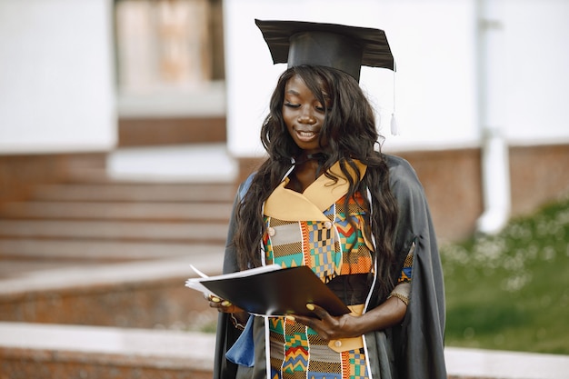 Free photo young afro american female student dressed in black graduation gown. campus as a background