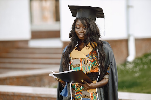 Young afro american female student dressed in black graduation gown. Campus as a background