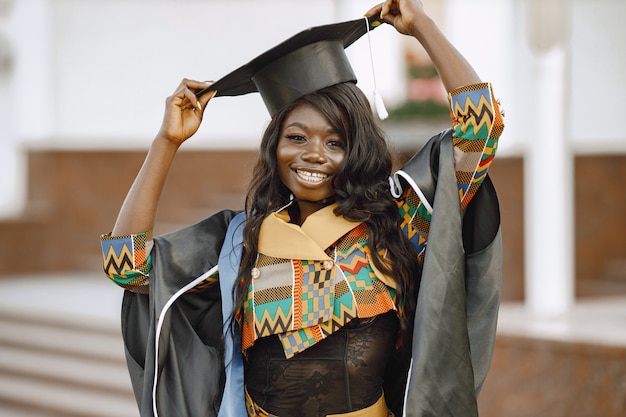 Young afro american female student dressed in black graduation gown. Campus as a background