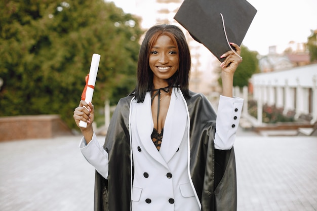 Young afro american female student dressed in black graduation gown. campus as a background