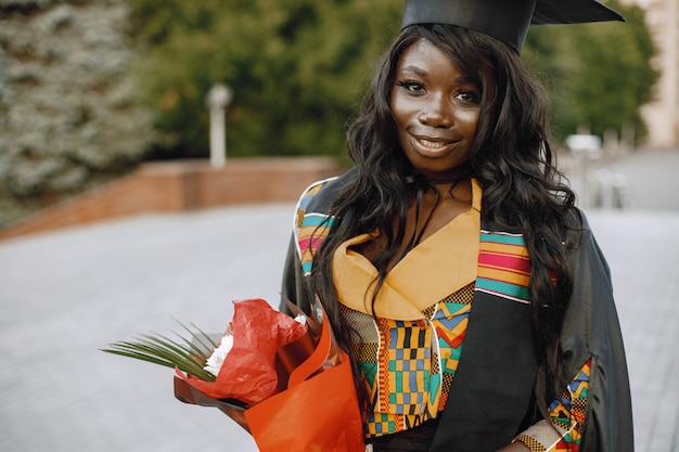 Young afro american female student dressed in black graduation gown. Girl posing for a photo and holding a flowers