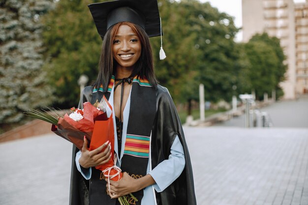 黒の卒業式のガウンに身を包んだ若いアフリカ系アメリカ人の女子学生。写真のポーズをとって花を持っている女の子