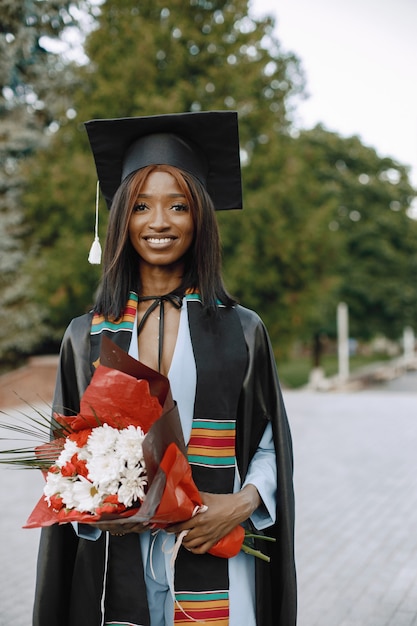 검은 졸업 가운을 입은 젊은 아프리카계 미국인 여학생. 사진을 위해 포즈를 취하고 꽃을 들고 소녀