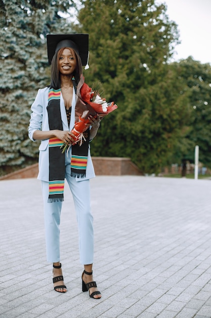 青い服とアカデミックキャップを着た若いアフリカ系アメリカ人の女子学生。写真のポーズをとって花を持っている女の子