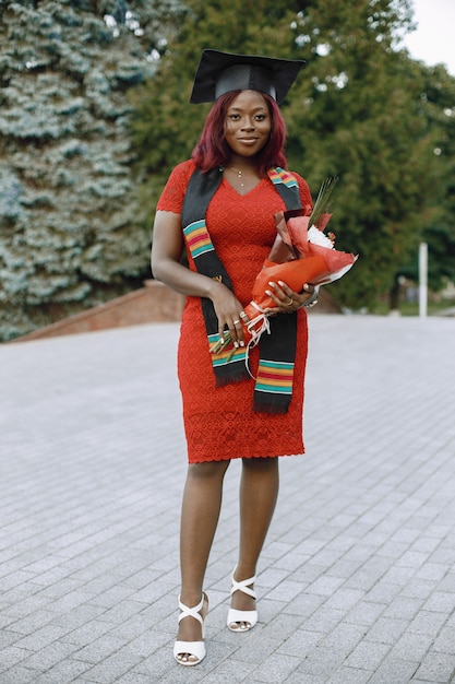 Молодая афро-американская студентка, одетая в красное платье и академическую кепку. Девушка позирует для фото и держит цветы