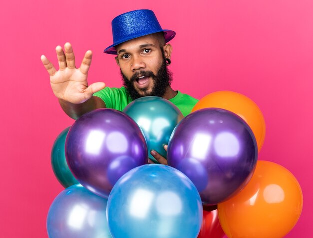 Young afro-american guy wearing party hat standing behind balloons isolated on pink wall