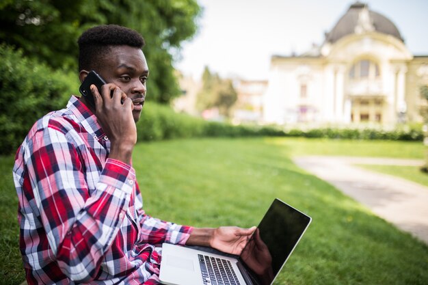 Молодой афроамериканец разговаривает по телефону, сидя на зеленой траве с ноутбуком на городской улице