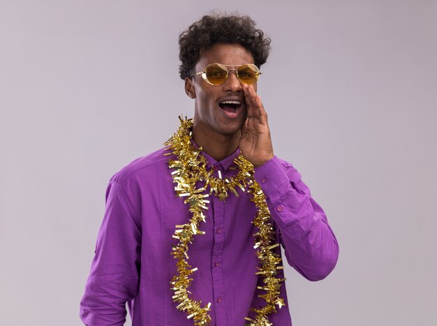 Young afro-american man wearing glasses with tinsel garland around neck looking at camera whispering isolated on white background