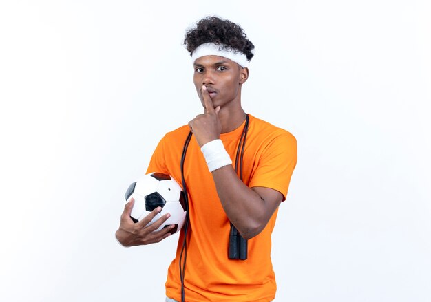 молодой афро-американский спортивный мужчина с головной повязкой и браслетом держит мяч, показывая жест тишины со скакалкой на плече
