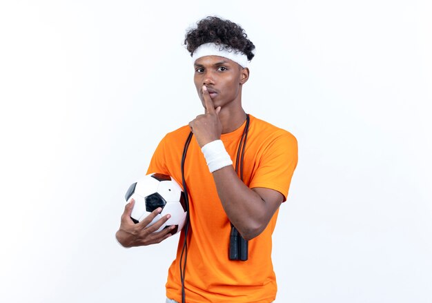 young afro-american sporty man wearing headband and wristband holding ball showing silence gesture with jump rope on shoulder 