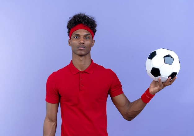 young afro-american sporty man wearing headband and wristband holding out ball to side isolated on blue wall