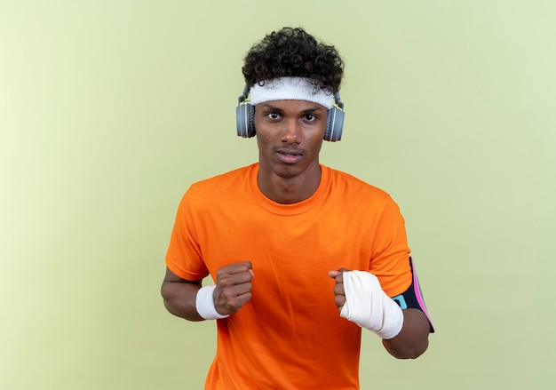 Giovane uomo sportivo afro-americano che indossa la fascia e il braccialetto e la fascia da braccio del telefono con le cuffie in piedi nella posa di combattimento isolata sulla parete verde