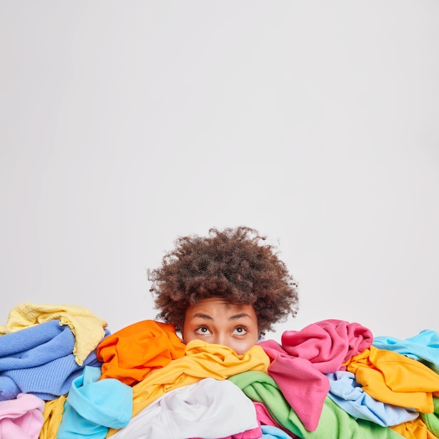 Young Afro American woman surrounded by different colorful clothes sorts out wardrobe focused above isolated over white wall blank space for your advertising content. Nothing to wear concept
