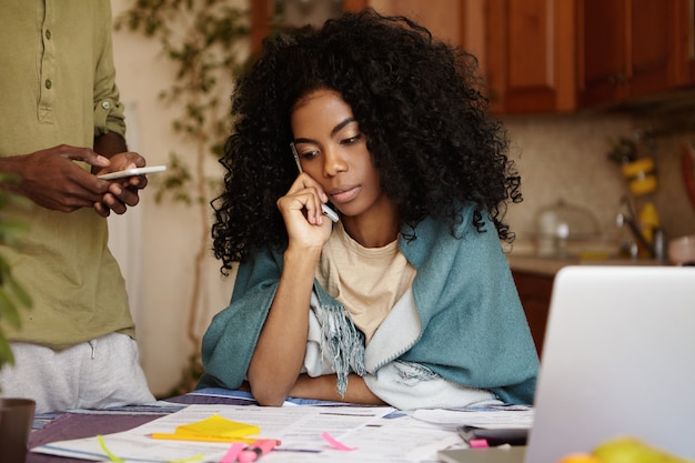 ラップトップと紙でテーブルに座って、ローンの借金について情報提供する銀行と携帯電話で話しているキッチンでの財政を通じて働いている間心配している巻き毛の若いアフリカ系アメリカ人女性