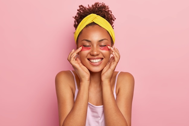 若いアフロの女性モデルは、目の下にコラーゲンパッドを適用し、保湿治療を楽しんで、広く笑顔で、白い歯が新鮮で健康な肌を持っていることを示しています黄色のヘッドバンドを着用しています
