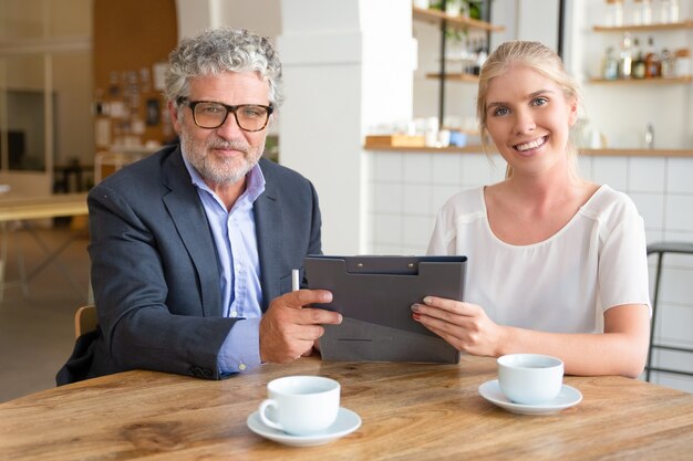 Young agent and mature customer meeting over cup of coffee at co-working, sitting at table, holding documents, 