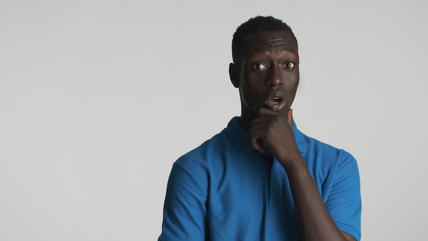 Молодой изумленный афроамериканец, задумчиво потирающий подбородок, выглядит удивленным в камеру на белом фоне Вау выражение лица