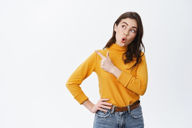 Young amazed girl say wow staring and pointing left at empty space showing advertisement with interested face expression standing against white background