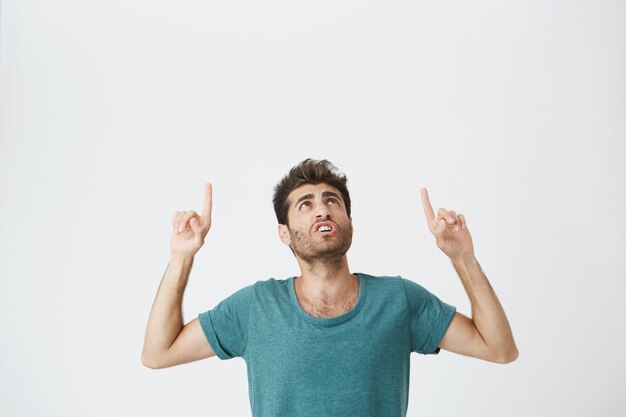 Young amazed stylish man with beard pointing his fingers at white wall with copy space for your advertisement or promotional information