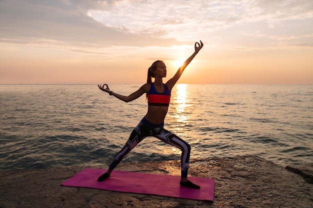 Молодая красивая женщина, осуществляющих на пляже, практикующих йогу в прекрасный восход солнца.
