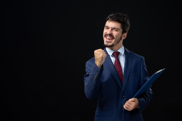 Young ambitious male office worker in suit holding documents and enjoying his succeess on isolated dark wall